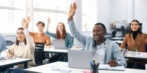 College-age students sitting in a classroom, raising their hands and smiling